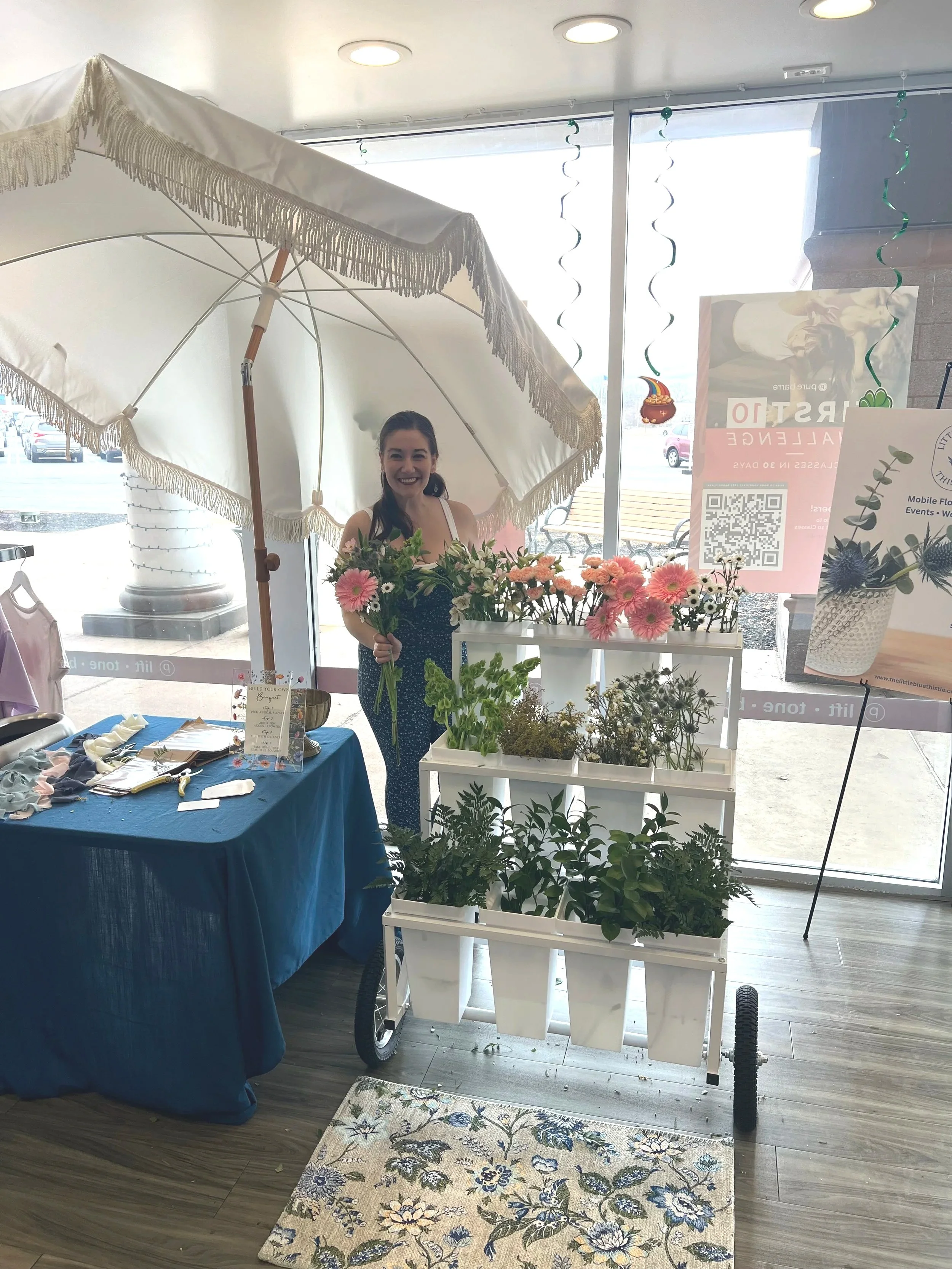 A woman standing behind a flower display cart with pink and white flowers, under a large white umbrella inside a store with glass windows.