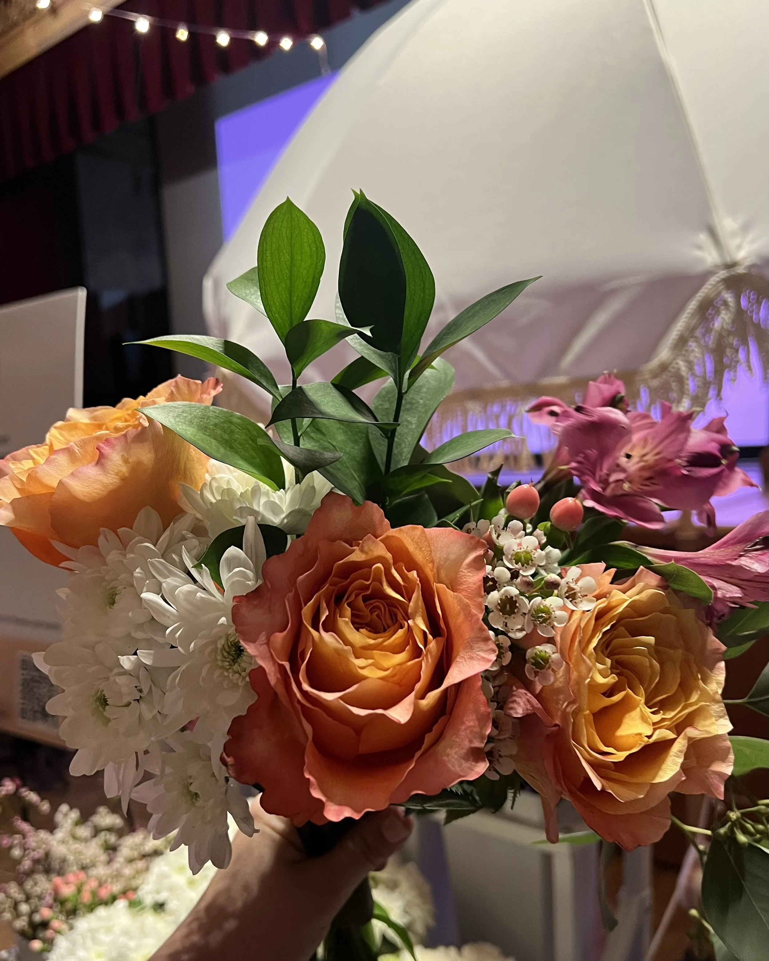 Close-up of a colorful flower bouquet with roses, daisies, and other flowers, held in a person's hand. In the background, there's a white decorative umbrella and stage curtains with string lights.