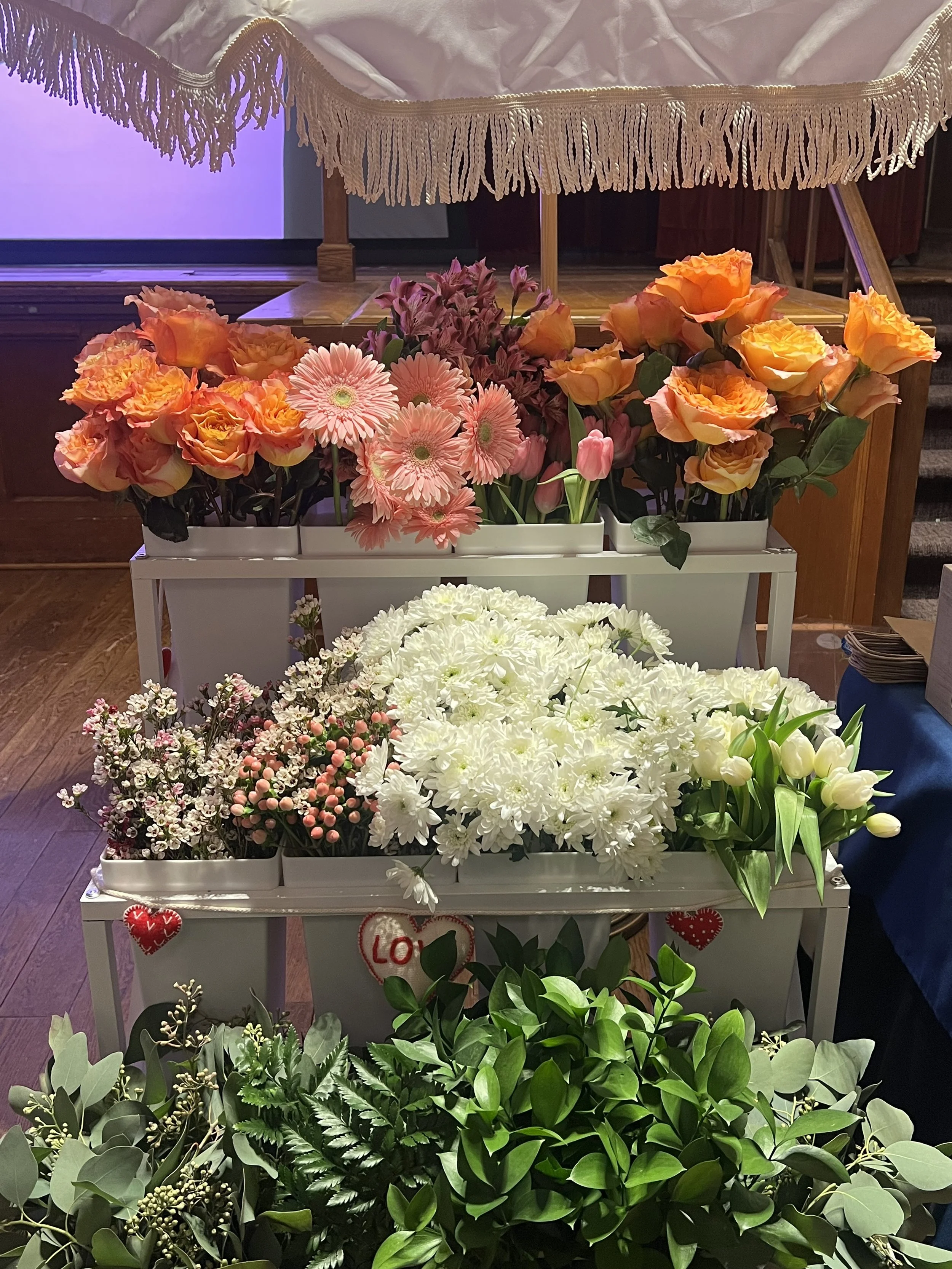Display of various colorful flowers, including roses, tulips, daisies, and filler flowers, arranged on white tiered shelves with valentine's decorations, set in an indoor setting.