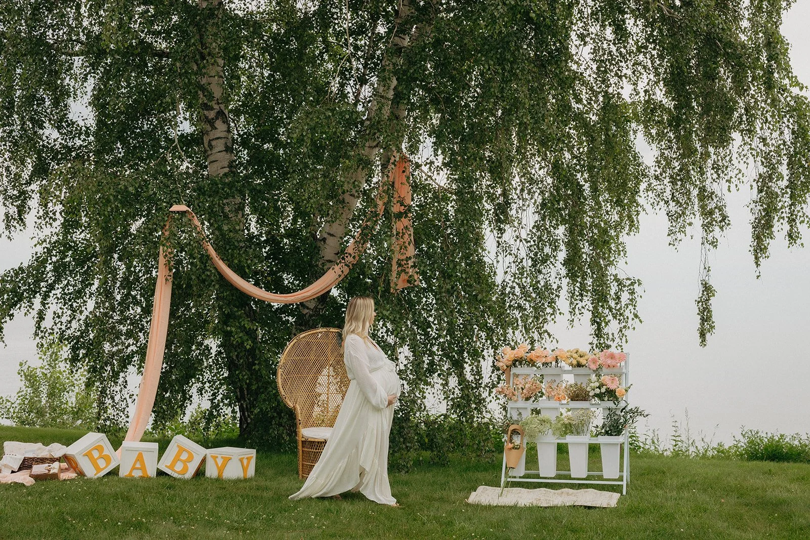 A pregnant woman in a white dress standing outside under a large tree, with decorations including a pink drape, a wicker chair, floral arrangement, and blocks spelling 'BABY' on the grass.