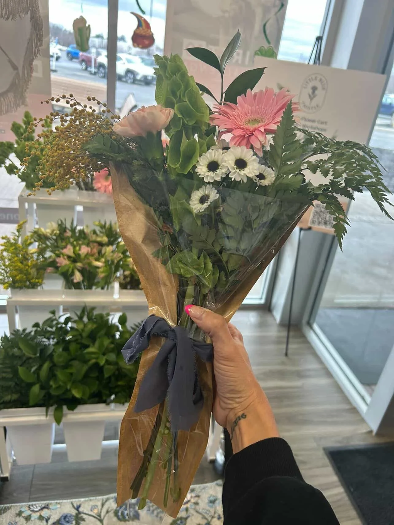 A person holding a bouquet of mixed flowers wrapped in brown paper and tied with a dark gray ribbon, inside a flower shop.