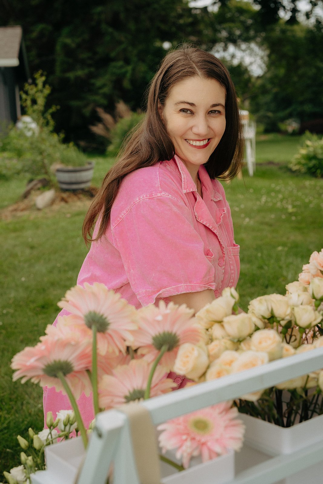 A woman with long brown hair and a bright smile, wearing a pink shirt, stands outdoors surrounded by flowers and greenery.