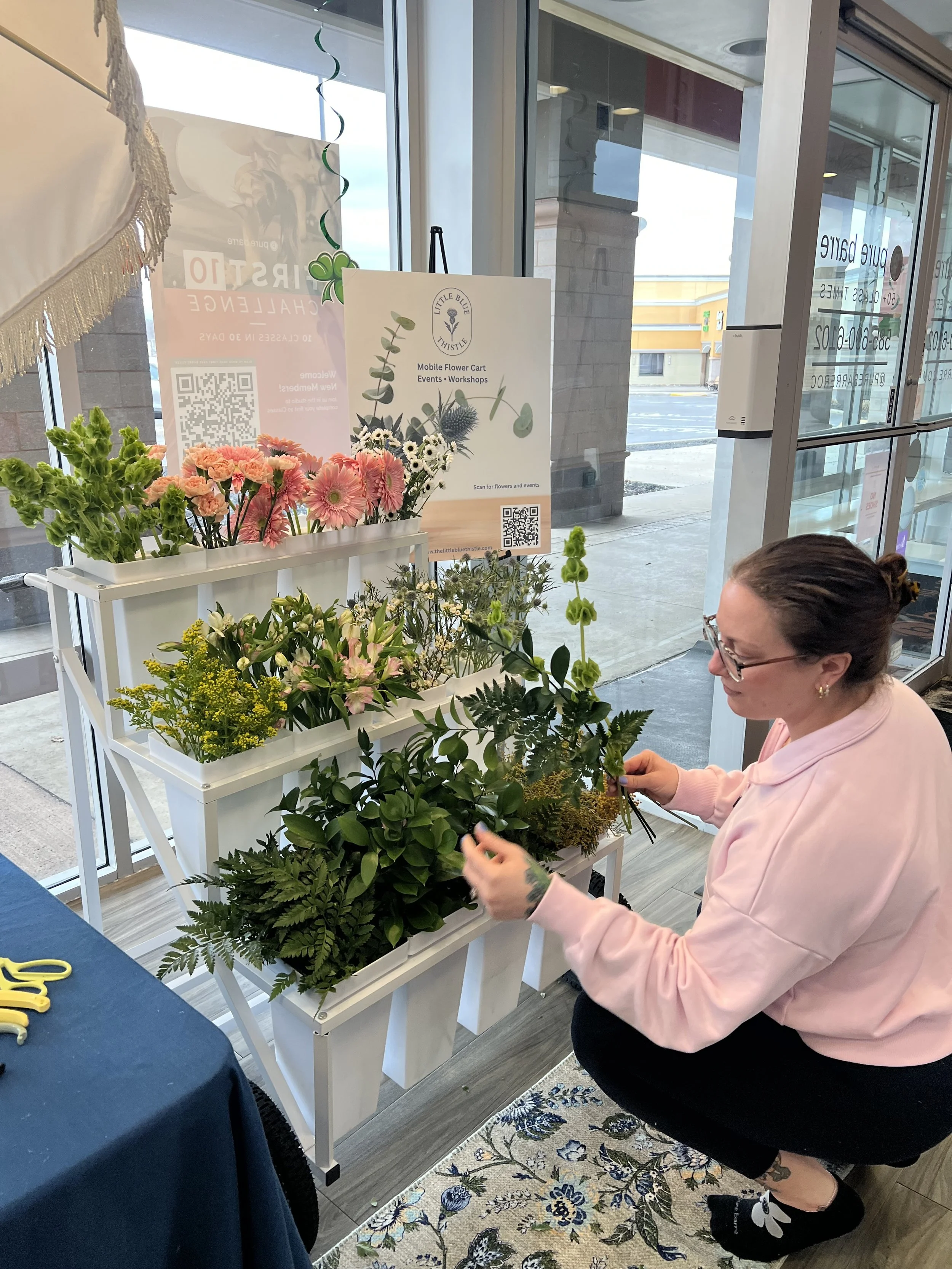 A woman in a pink sweatshirt arranging flowers on a white display stand inside a store. The stand has multiple trays of colorful flowers, including pink, white, and yellow blossoms. A sign for Little Blue Thistle with QR codes is on the display, and 