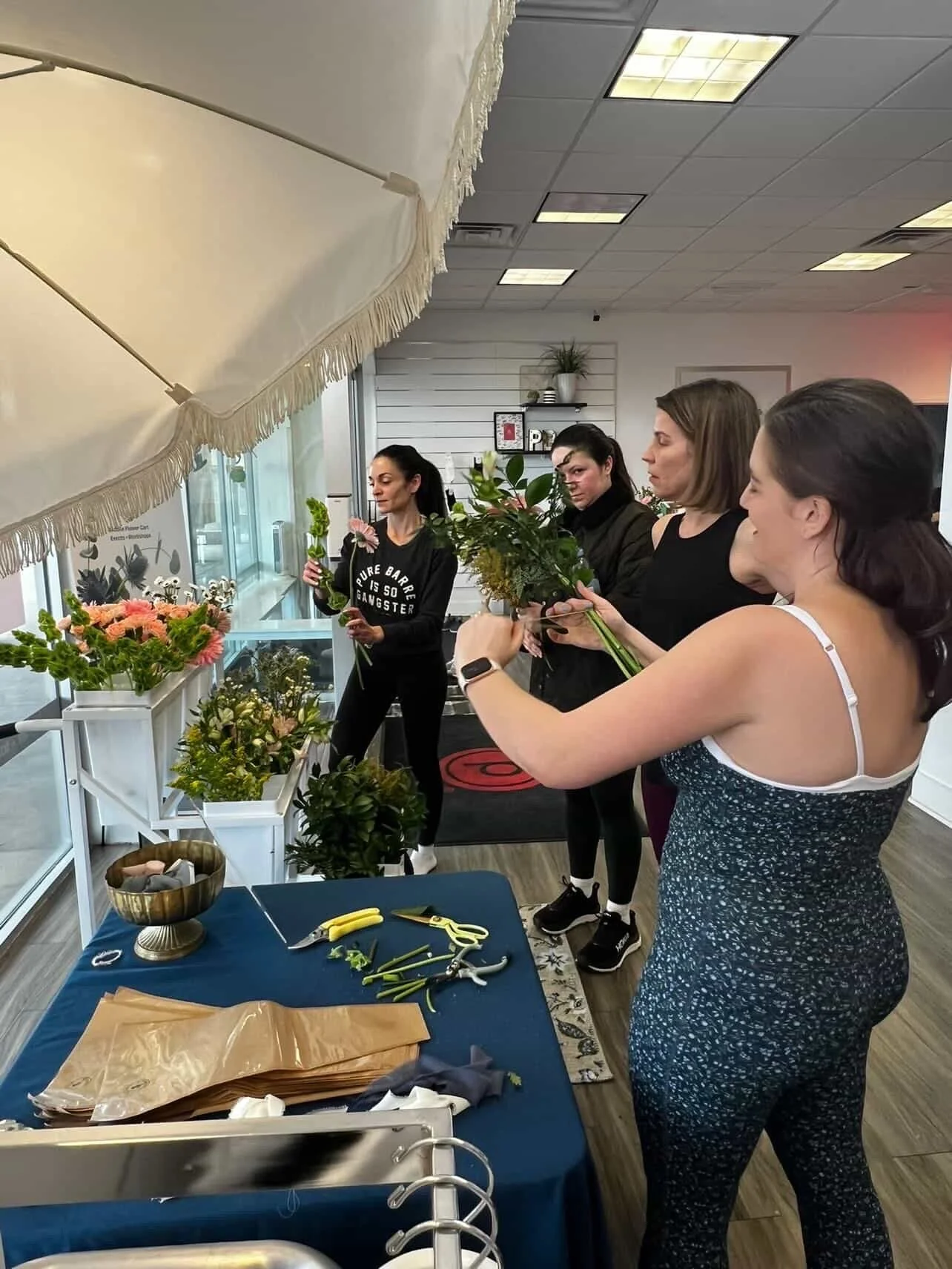 Four women are gathered around a table with flowers and gardening tools, preparing flower arrangements in a bright indoor space.