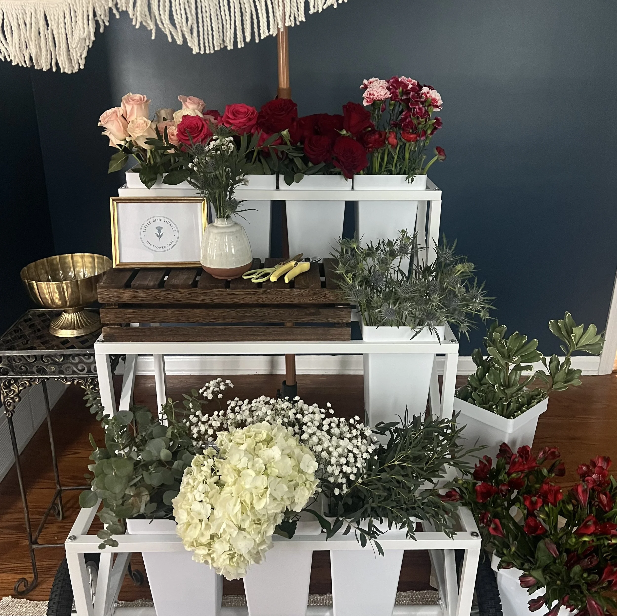 Various flowers including roses, hydrangeas, and anthuriums arranged on white and wooden shelves with a dark blue wall in the background.