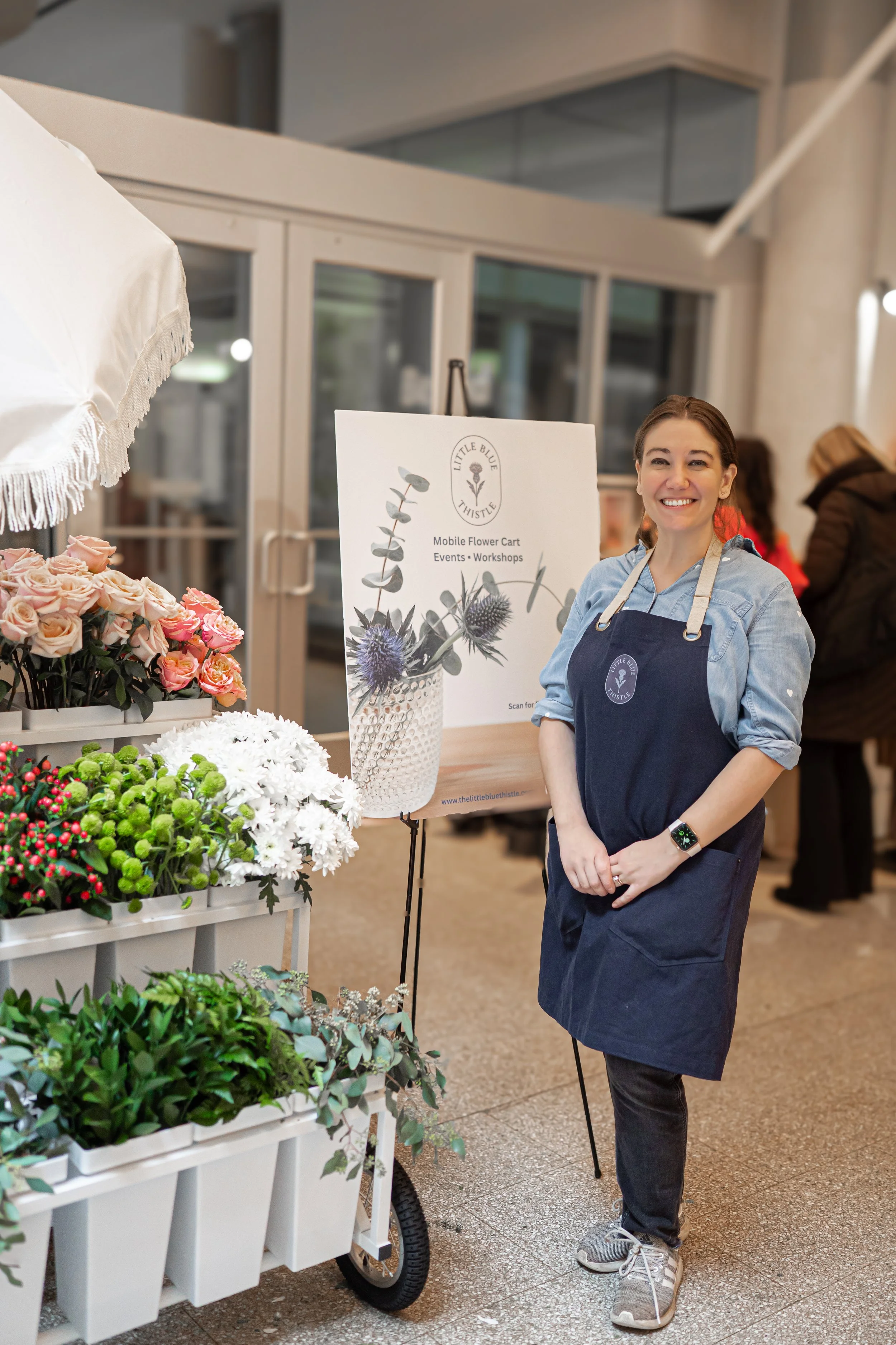 Founder of Little Blue Thistle standing beside her mobile flower cart at an indoor event, surrounded by fresh blooms and floral signage.