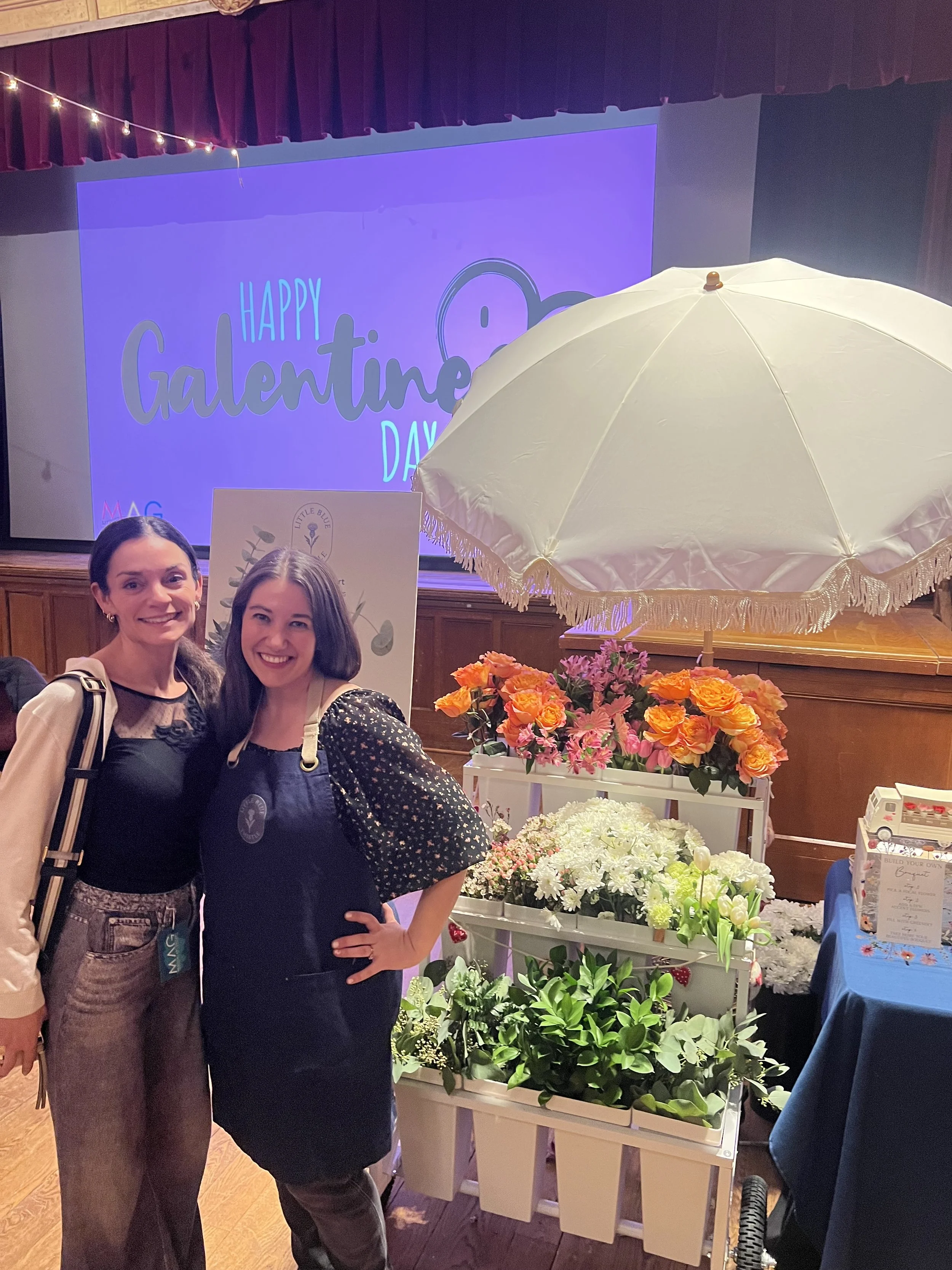 Two women smiling at a Valentine's Day event standing in front of a display with flowers and a vintage white umbrella, with a screen in the background reading 'Happy Valentine's Day'.