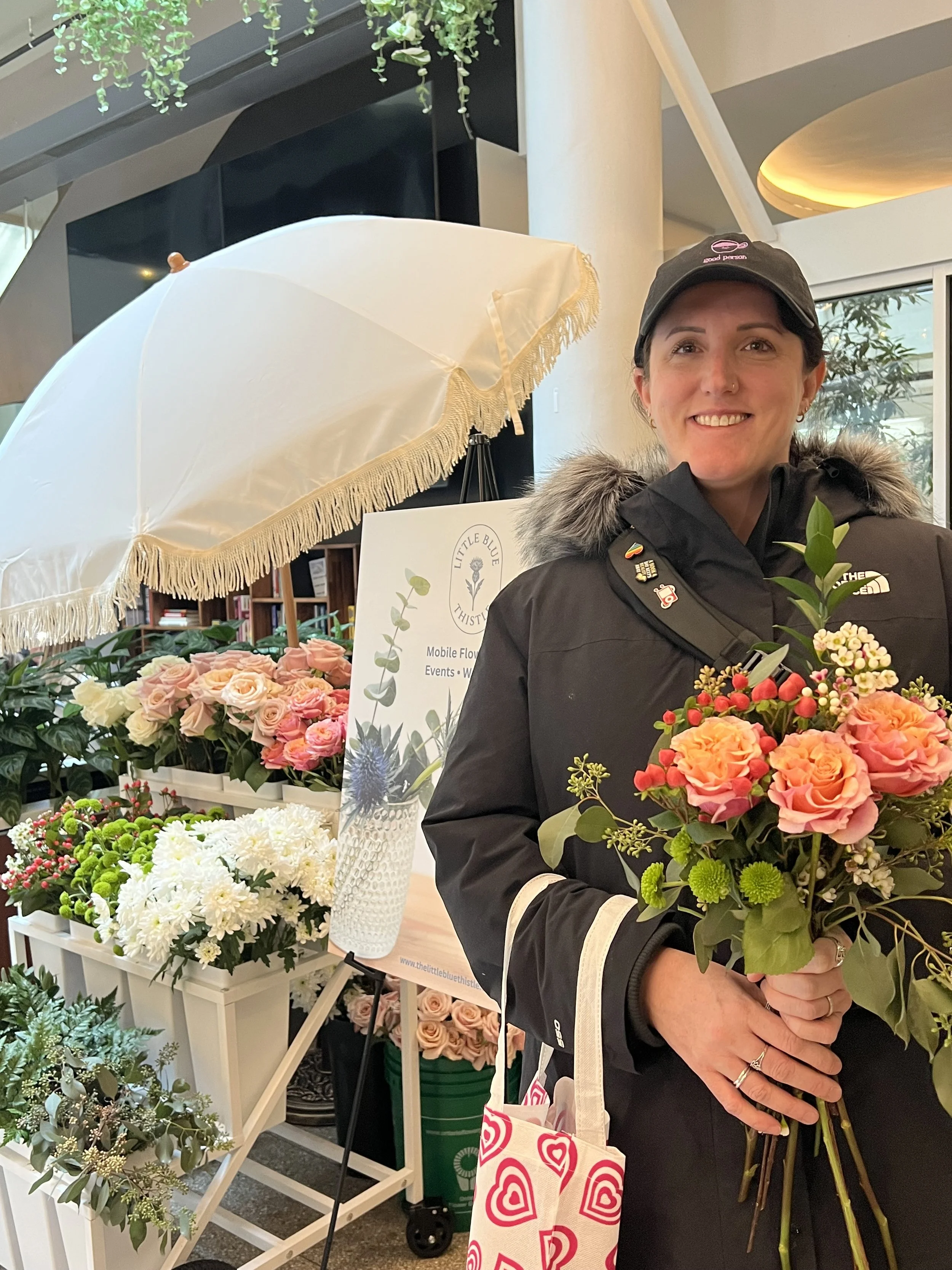 A smiling woman wearing a black jacket and a black cap holding a bouquet of pink roses and other flowers in a flower shop or display area with white and pink flowers, a white umbrella, and a sign with a logo and plant graphics.