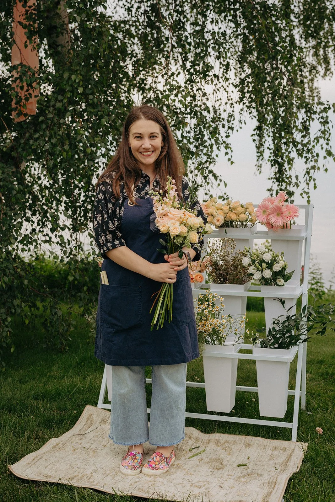 A woman standing outdoors, holding a bouquet of light pink and cream roses, smiling. Behind her is a display of various flower arrangements on white shelves, and trees in the background.