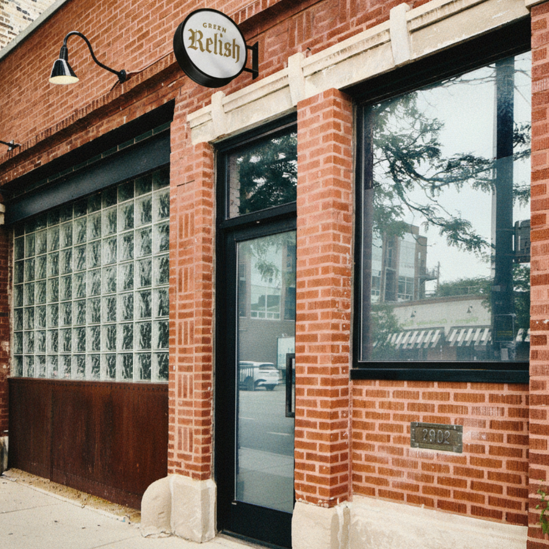 Sign for Green Relish restaurant mounted above the entrance to a brick building with a glass door and large window.
