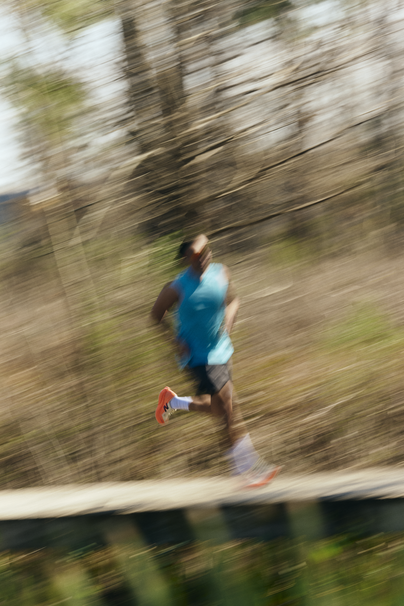 A man running outdoors on a trail, wearing a blue sleeveless shirt, black shorts, orange running shoes, and white socks, with a blurred background of trees and bushes indicating motion.