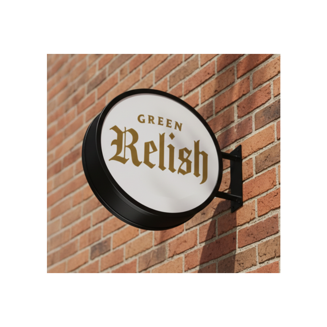 Round sign with black frame reading "Green Relish" in gold letters, mounted on a brick wall.