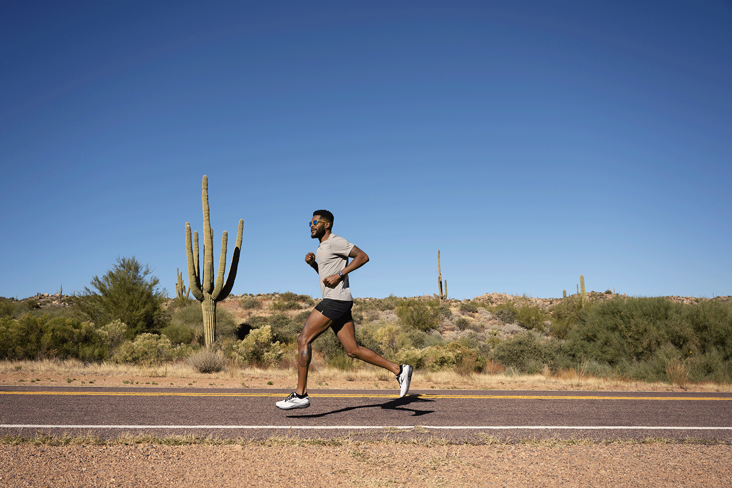 Man running on a paved desert road with cacti and desert shrubs under a clear blue sky.