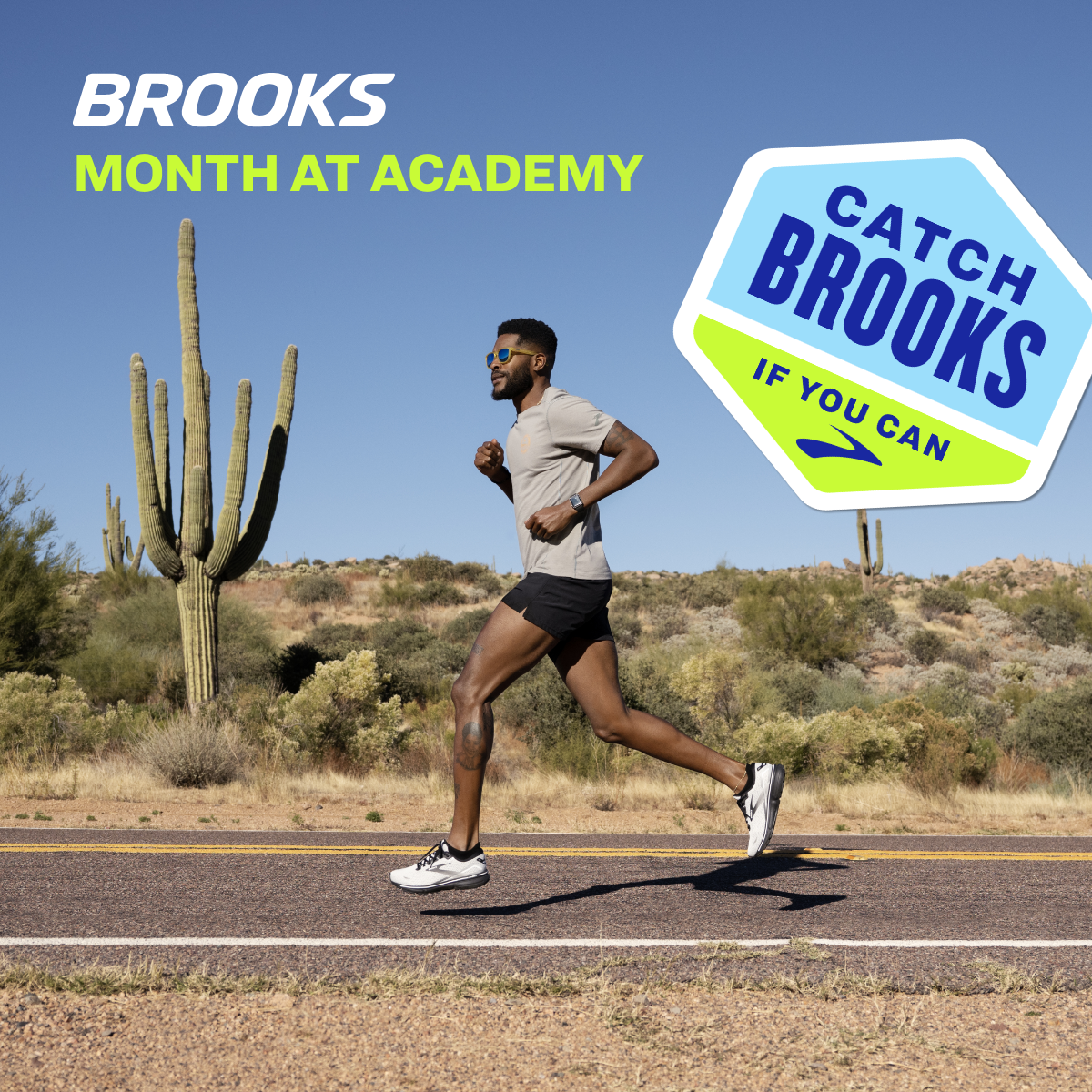 A man jogging on a desert road with cacti and bushes, wearing sunglasses, a light-colored t-shirt, black shorts, and running shoes, under a clear blue sky.