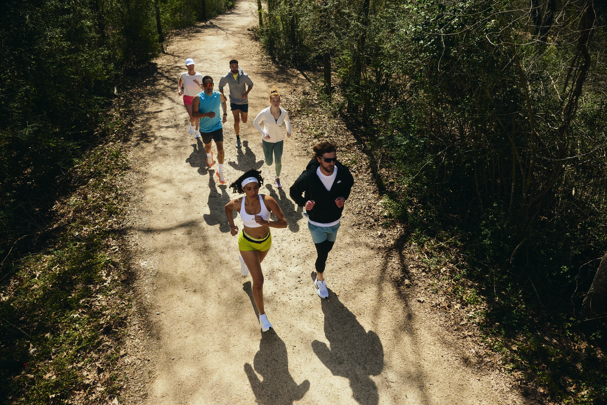 A group of seven people running on a dirt trail through a wooded area.