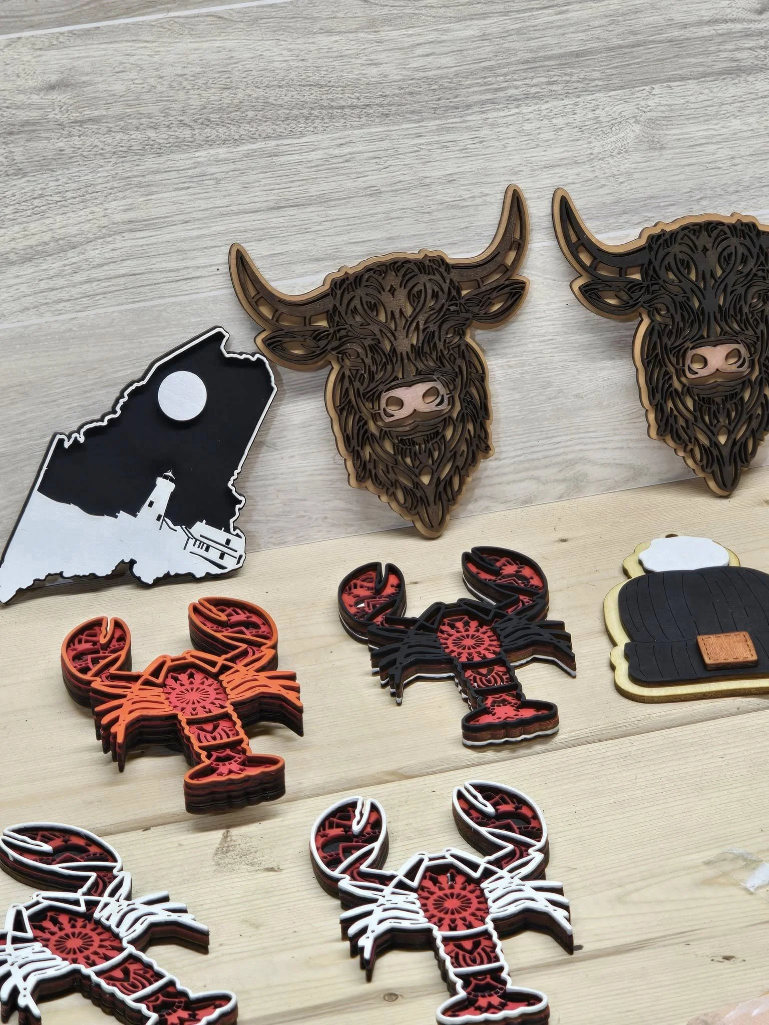 Various decorative items including a black and white shape of Maine, two large animal head wall art pieces resembling highland cows, and several lobster-shaped stickers or patches in red, white, and black on a light wooden surface.