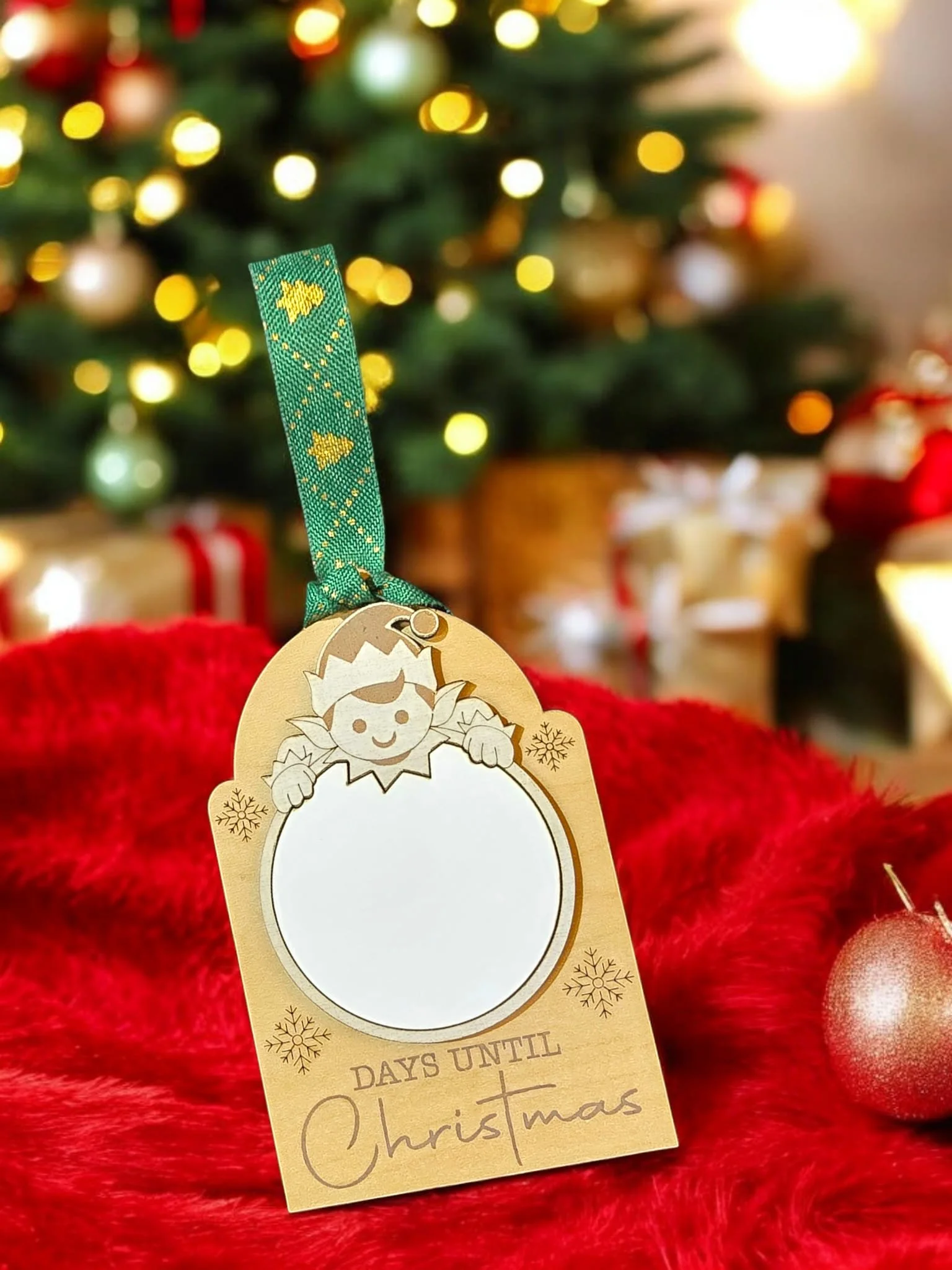 A Christmas-themed countdown tag with a cartoon elf peeking over a blank circle, decorated with snowflakes and the text "DAYS UNTIL Christmas" on a red furry surface, with a decorated Christmas tree and presents in the background.
