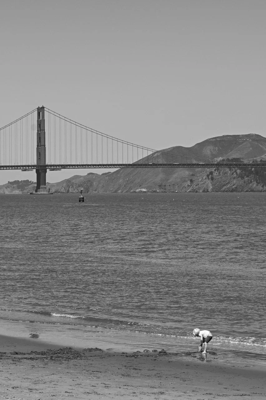 A young child playing near the shoreline on a beach with the Golden Gate Bridge and hills in the background, black and white photograph.