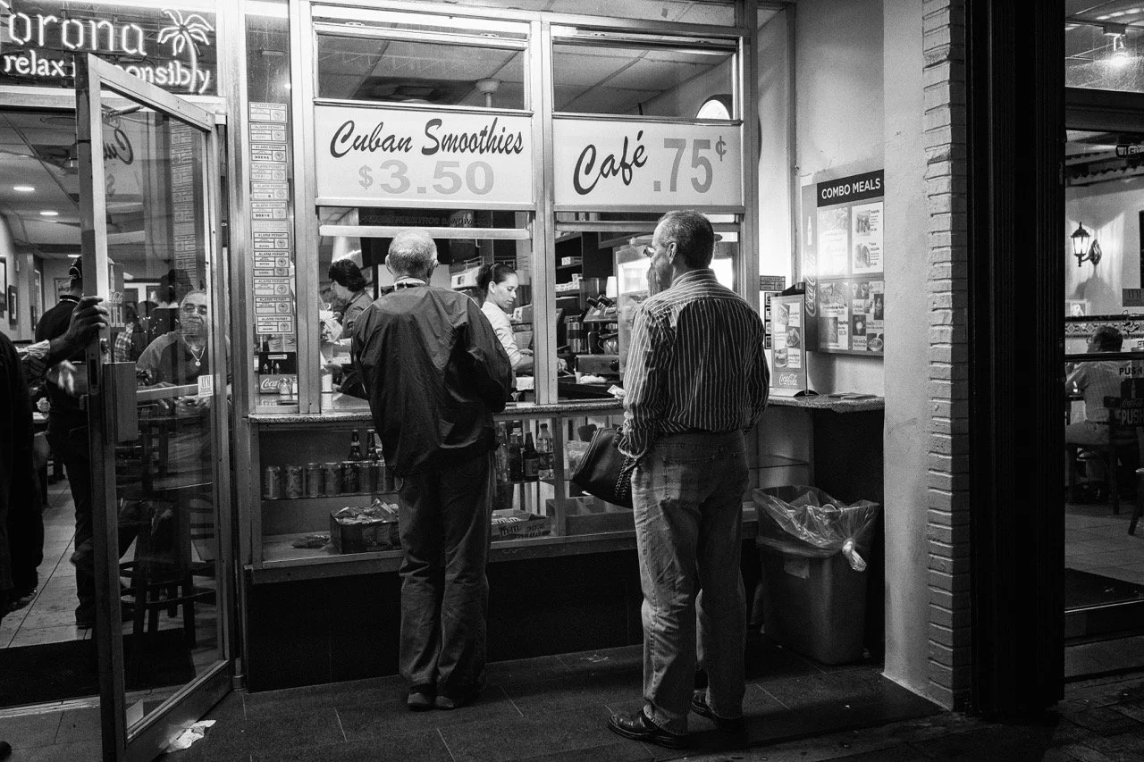 People standing at a Cuban smoothie and café stand inside a restaurant, with signs advertising Cuban smoothies for $3.50 and coffee for 75 cents, and hamburgers and combo meals on a menu.