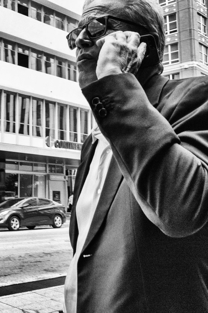 A man in business attire, wearing sunglasses, talking on a cellphone outside an urban building.