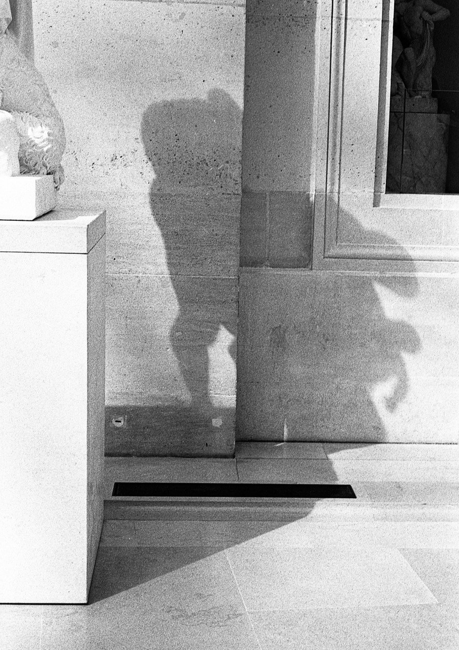 Black and white photo showing the shadows of two people cast on a wall and a mirror, with one person wearing shorts and the other a hat.
