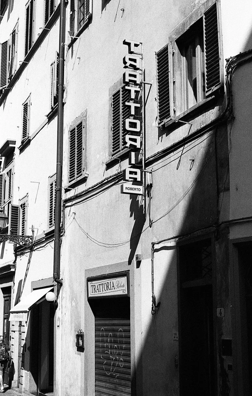 Black and white photo of a narrow street scene with a building that has multiple window shutters and signs. One vertical sign reads 'TRATTORIA' and another sign below it says 'Roberto.' Shadows cast on the wall suggest bright sunlight.