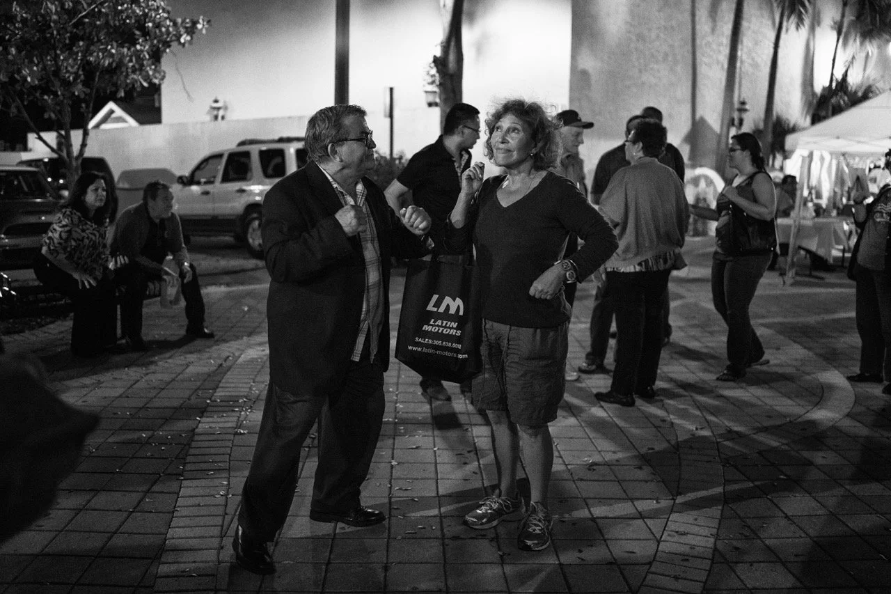 A black and white photo of two people having a conversation outdoors at night. The man on the left is wearing glasses, a blazer, and jeans. The woman on the right is holding a bag that reads "Latin Motors" and is dressed in a long-sleeve top and shor
