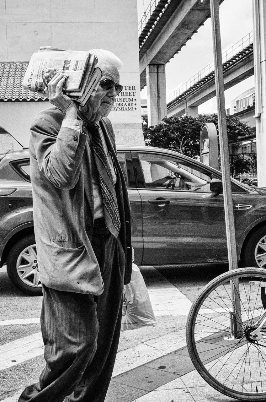 An elderly man wearing sunglasses and a suit jacket, holding a stack of newspapers to his ear as if talking on the phone, stands on a city street near parked cars and a bicycle.