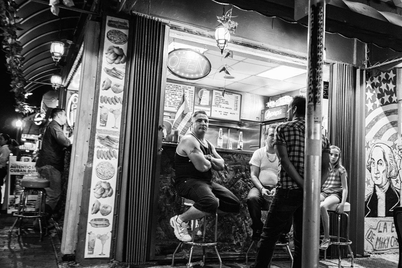 Black and white photo of a small restaurant or food stand with several people inside and outside. Inside, a man with tattoos and muscular build sits on a stool, crossing his arms. Two other men sit at a table, and a woman and young girl are seated on