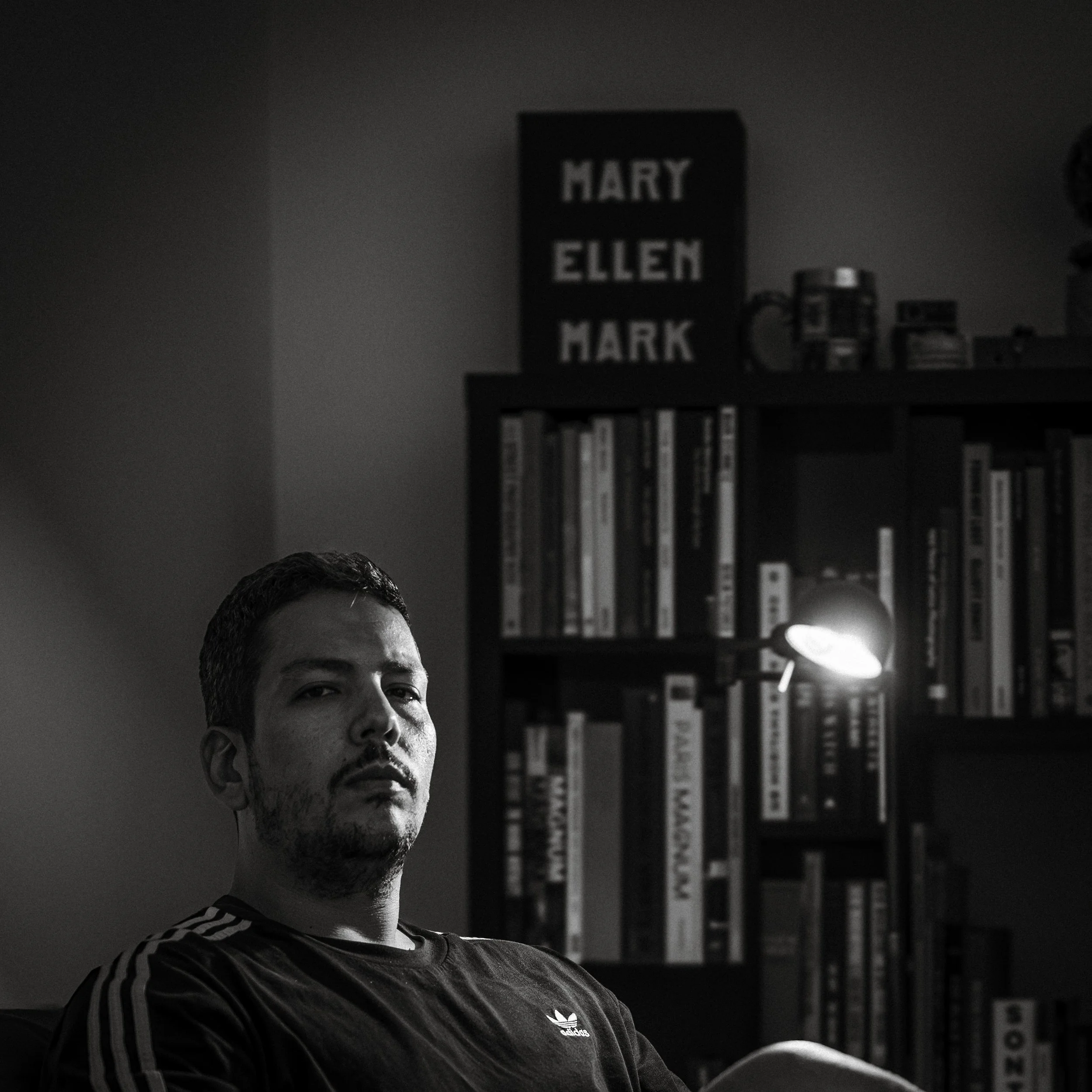A black and white photo of a man with short dark hair and a beard, sitting in a room with a bookshelf and a small lamp behind him.