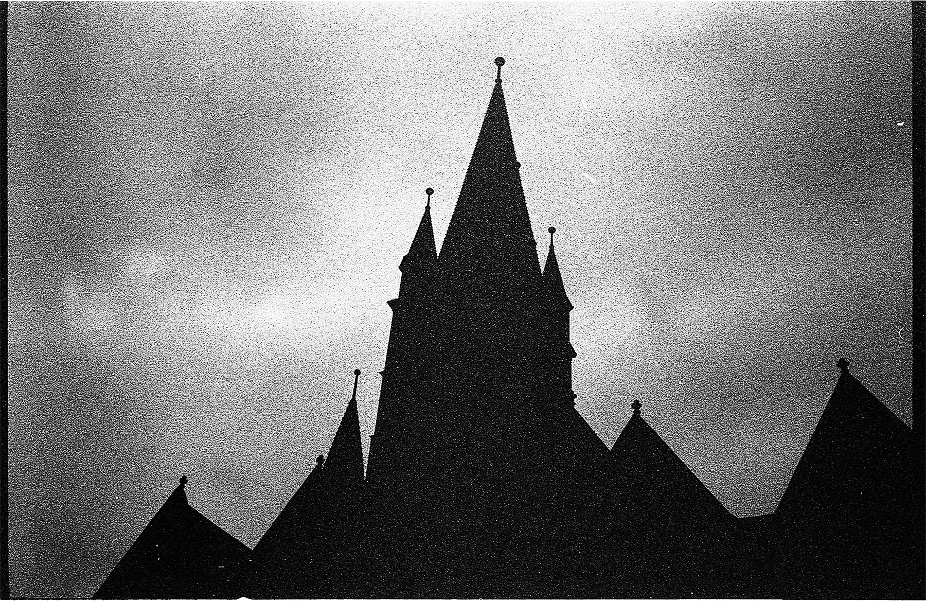 Black and white silhouette of a church with multiple tall, pointed spires against a cloudy sky.