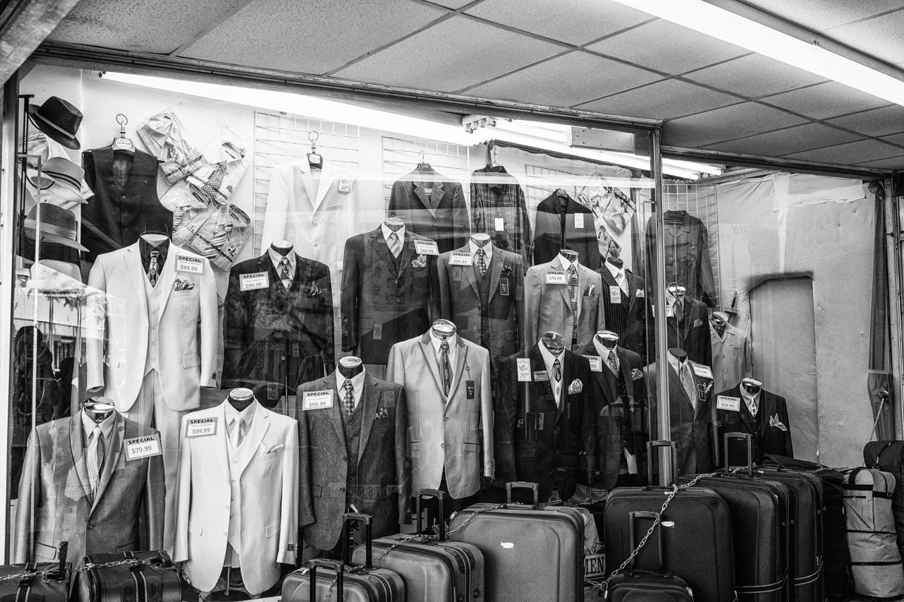 A storefront display of men's suits, ties, and accessories behind glass, with several suitcases in front.