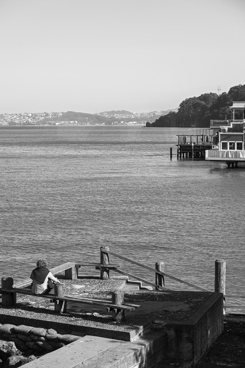 A black and white photo of a person sitting on a bench by the water, looking out at the bay with houses and trees on the opposite shore and some houses on stilts over the water.