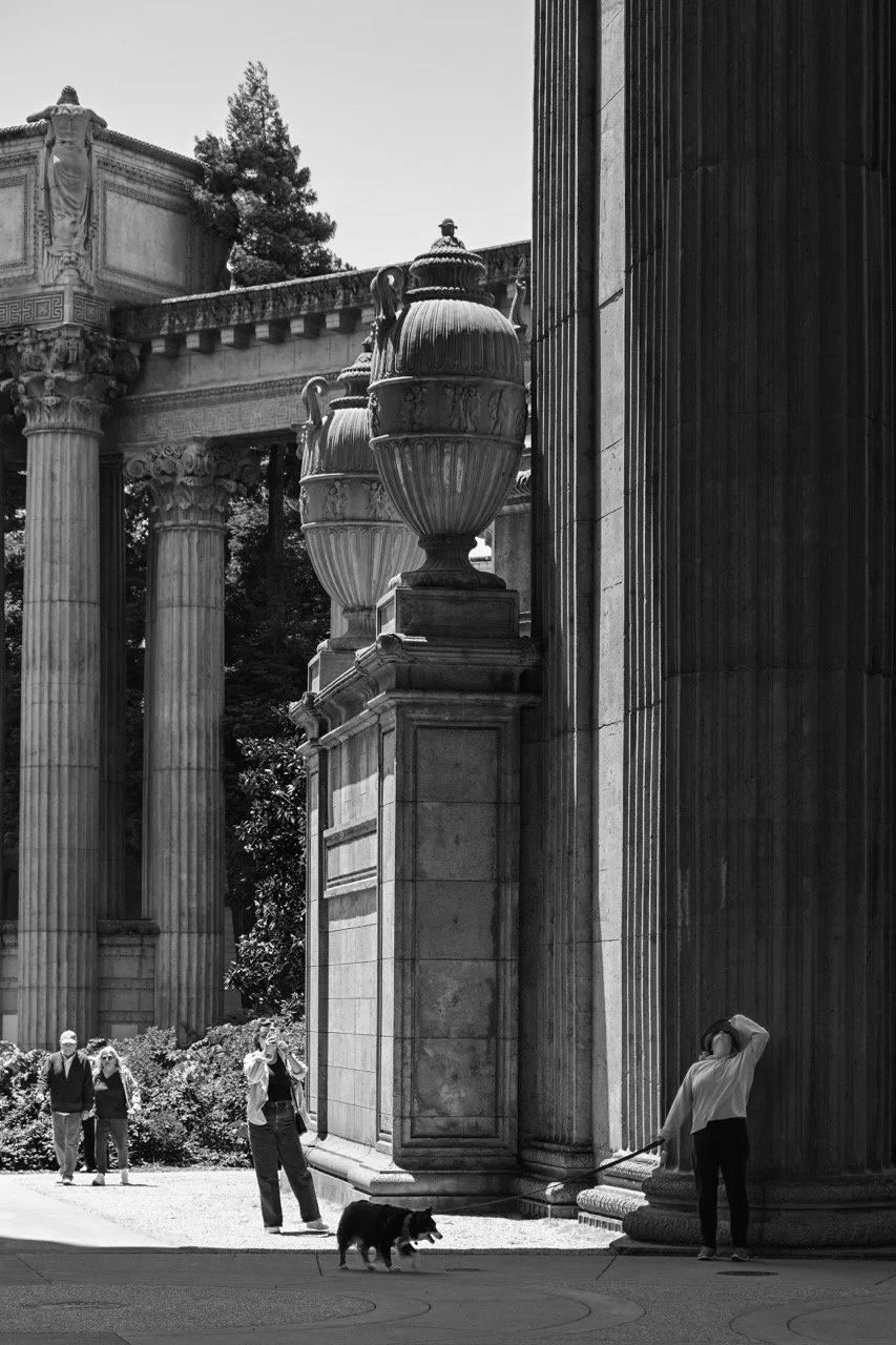 People walking near classical architectural columns and large decorative urns outdoors in black and white.