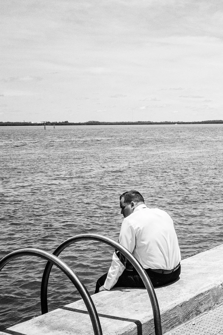 A man in business attire sitting alone at the edge of a waterfront, looking out over the water with a cloudy sky above.