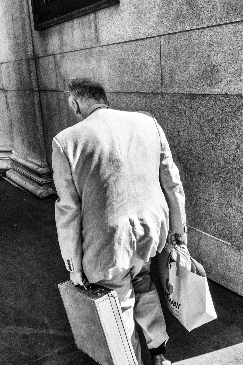 A man in a light-colored suit is walking on the sidewalk, carrying a briefcase in his right hand and a bag in his left hand, with his head bowed and leaning forward.