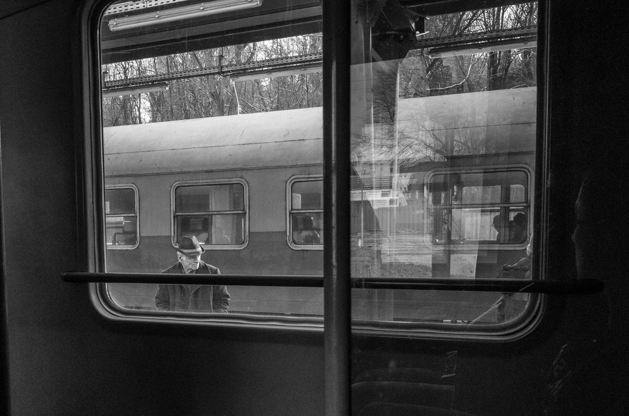 A black-and-white photo taken from inside a train, looking out the window at a man standing on the platform. The man is wearing a hat and coat, and is looking down, with a vintage train car on the platform behind him.