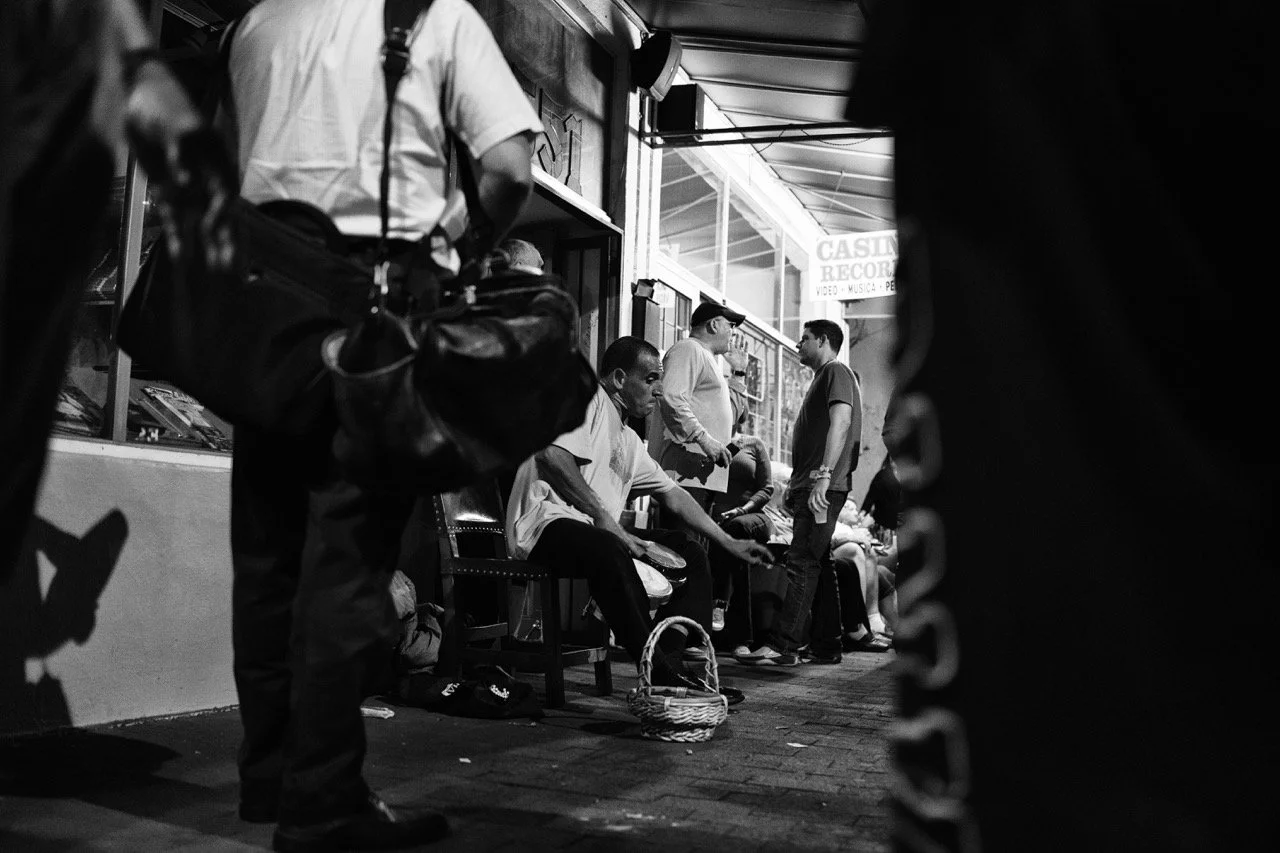 People sitting on benches outside a shop, with others standing and talking nearby, captured in black and white, with a focus on the seating area and passersby.