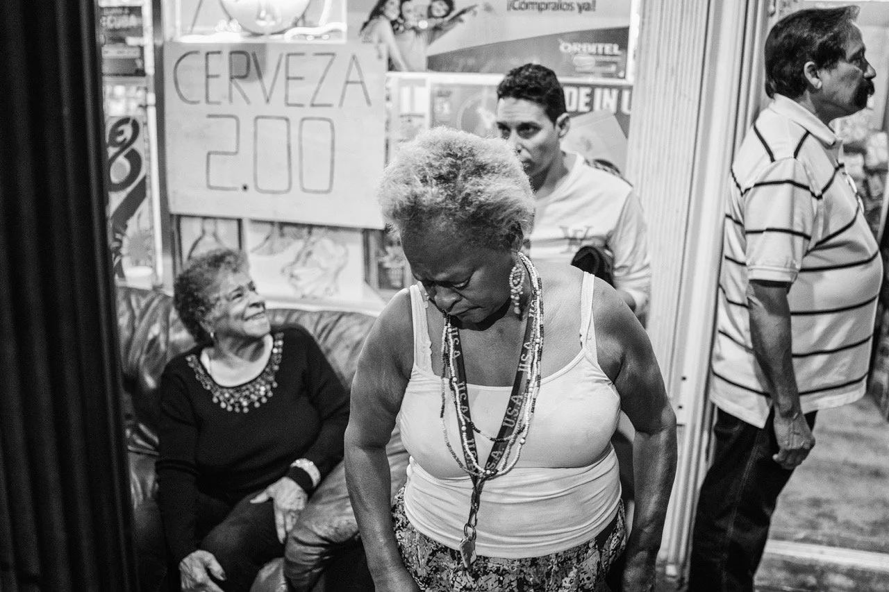 A black-and-white photo shows a woman with short, curly hair wearing a sleeveless top and jewelry, looking downward. Behind her, a woman is sitting on a couch, smiling and looking at her. Two men are standing nearby, one looking to the side and the o