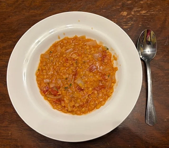 A white bowl filled with an orange-red soup. Lentils and chunks of tomato and onion are visible.