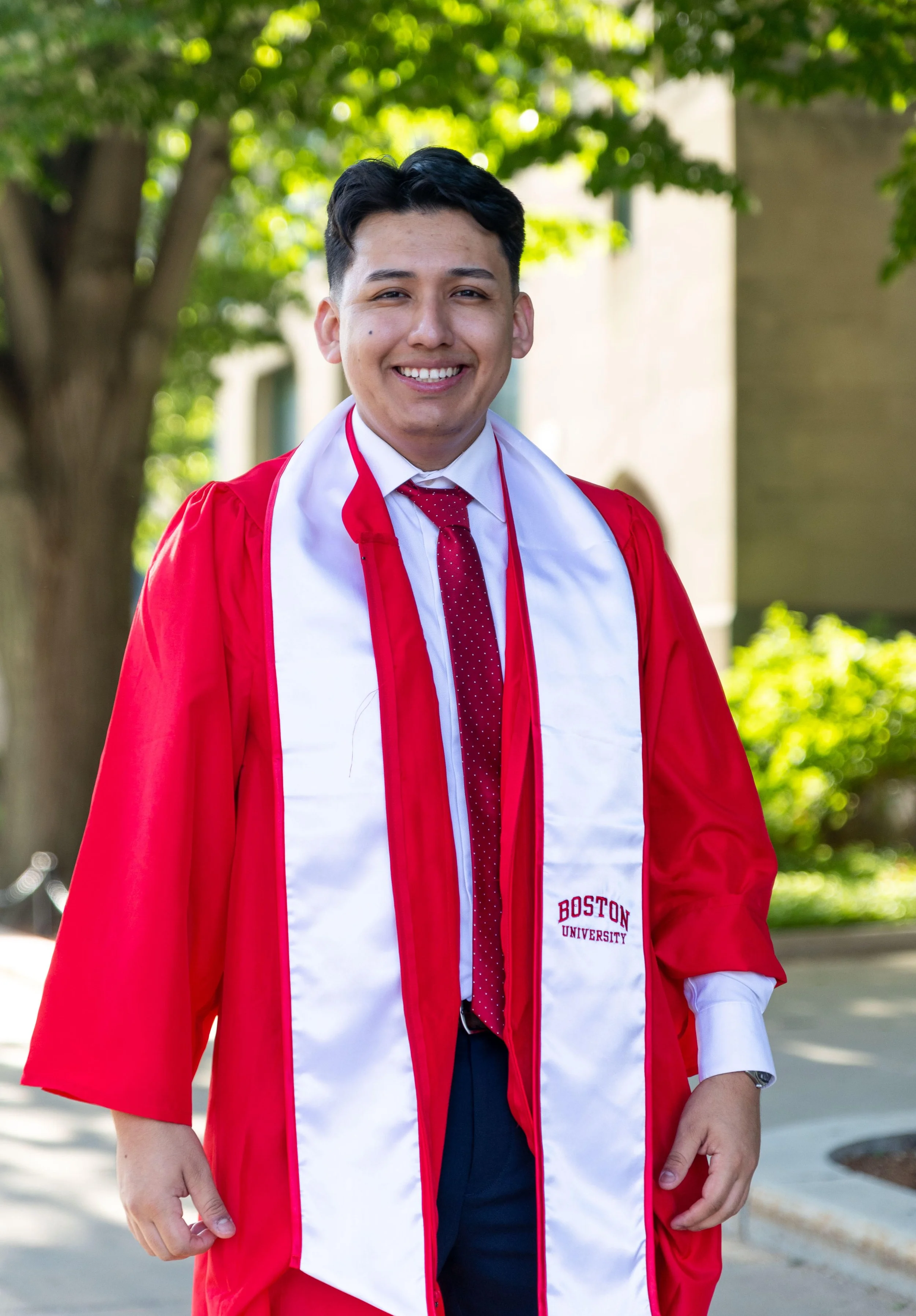 A young man in a red graduation gown and white stole, smiling outdoors with green trees and a building in the background.