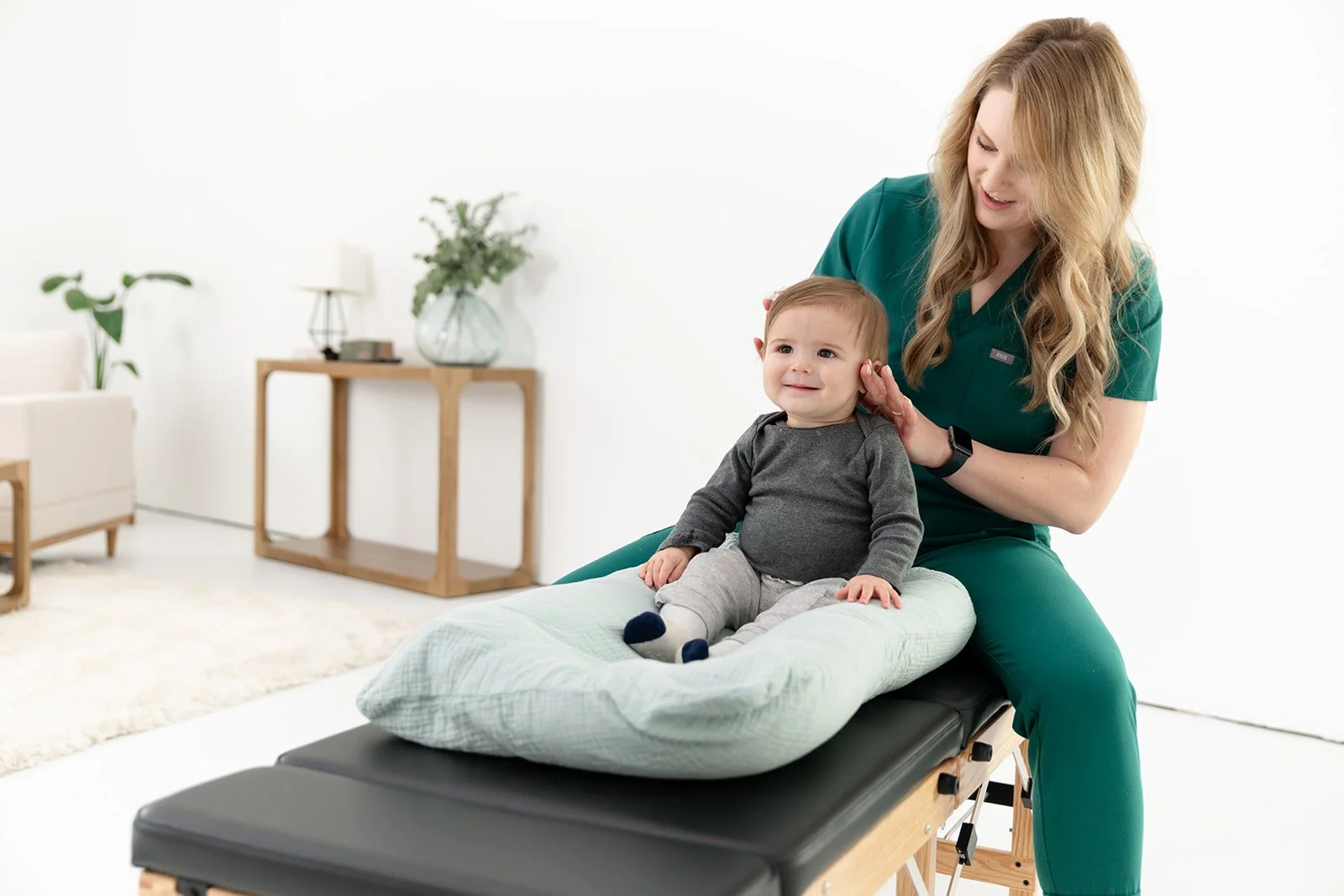 A female healthcare professional or therapist in green scrubs performing an ear examination on a young child lying on a padded table in a clinical setting.