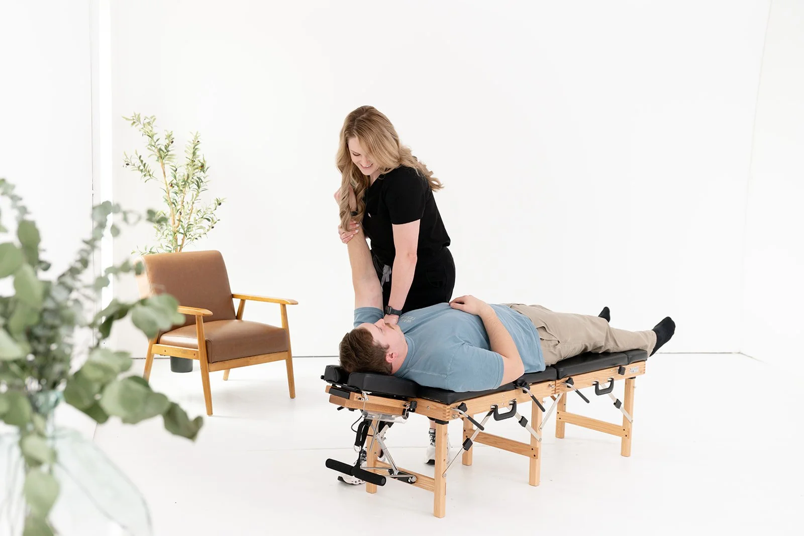 A woman in black assisting a man lying on a therapy table in a bright, white room.