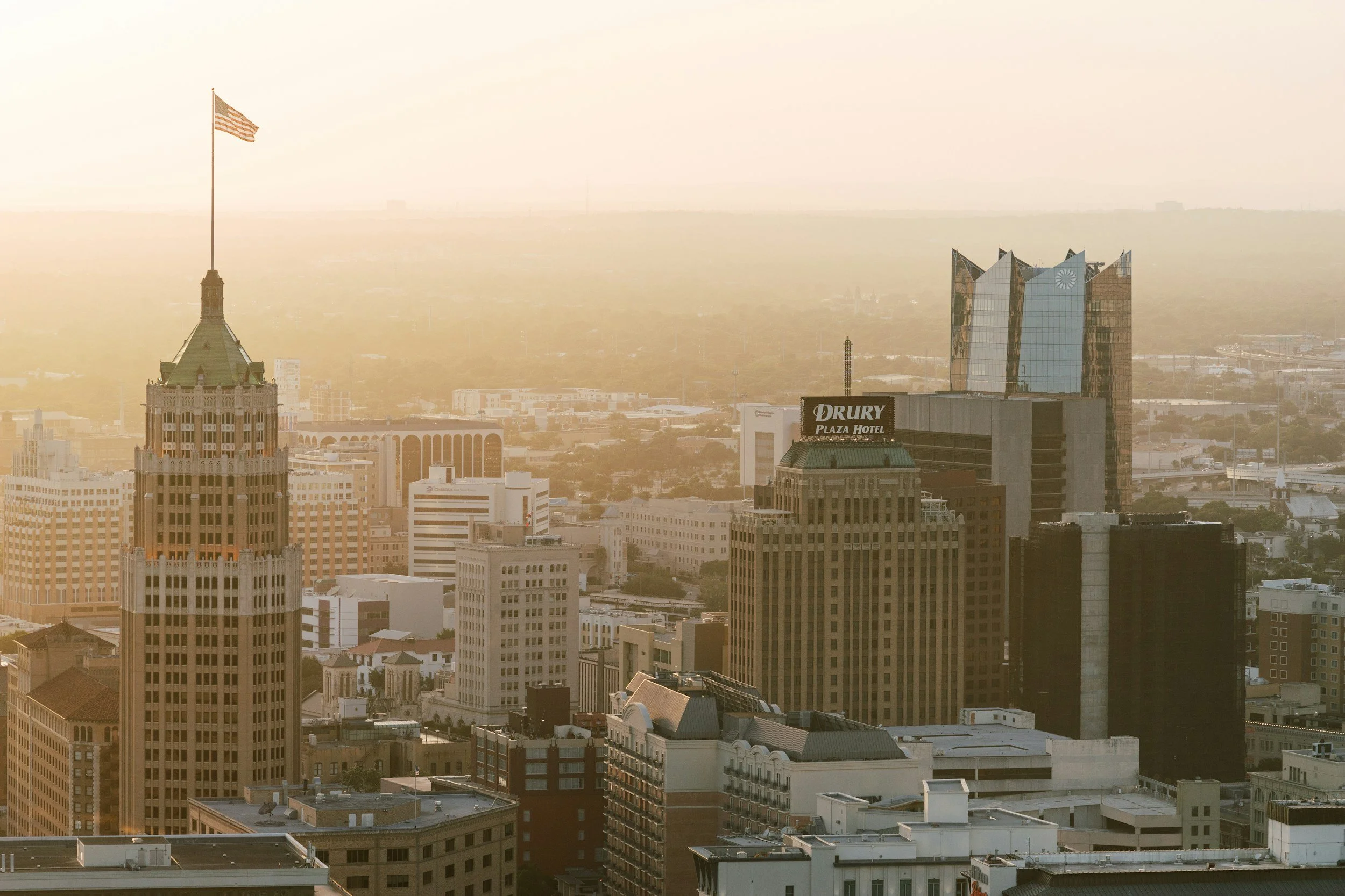 A city skyline with tall buildings in the foreground and a hazy background, including a prominent tower with an American flag on top. San Antonio, Texas