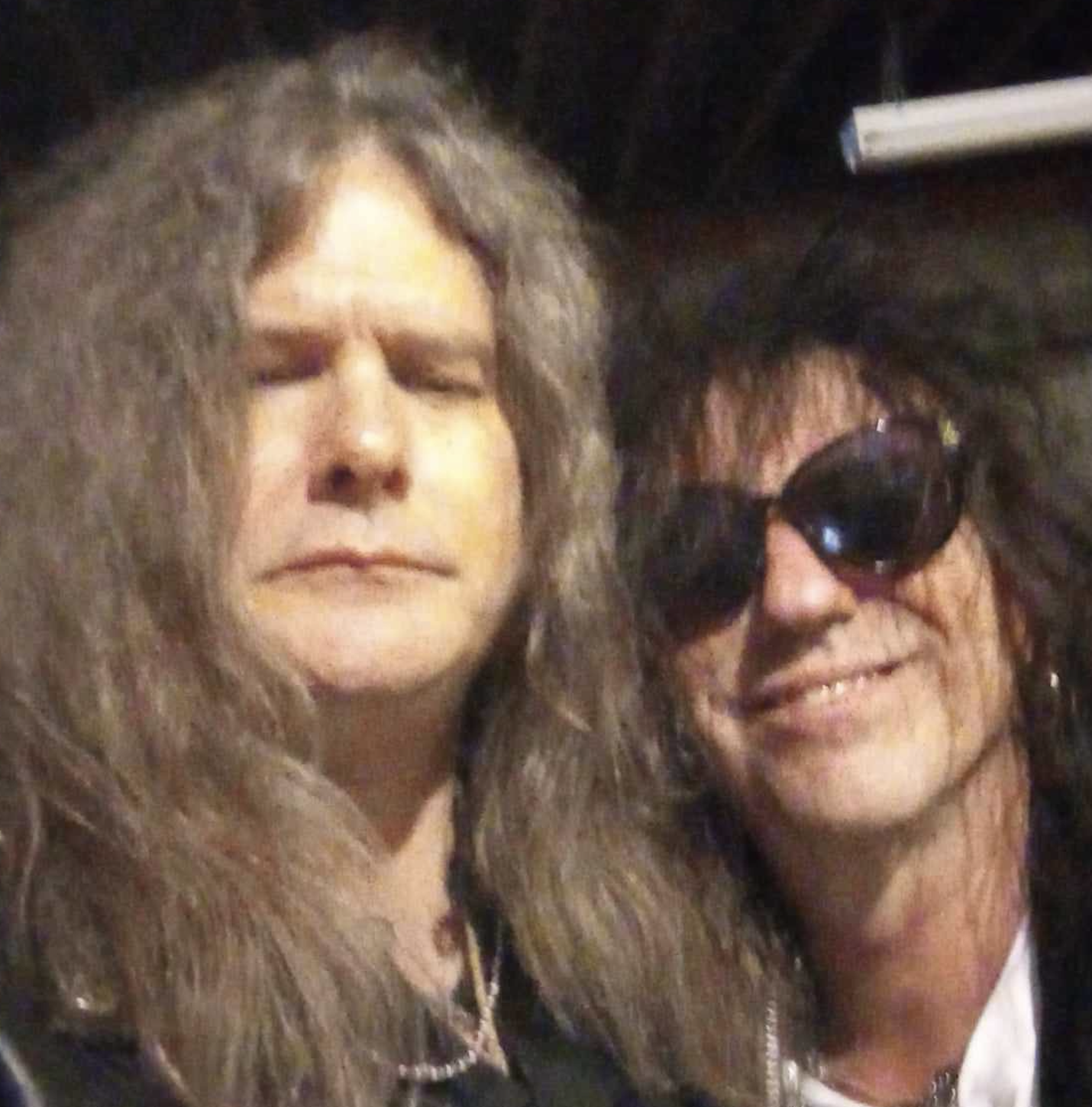 Two individuals with long, wavy hair, one wearing sunglasses and smiling, the other with eyes closed and a relaxed expression.