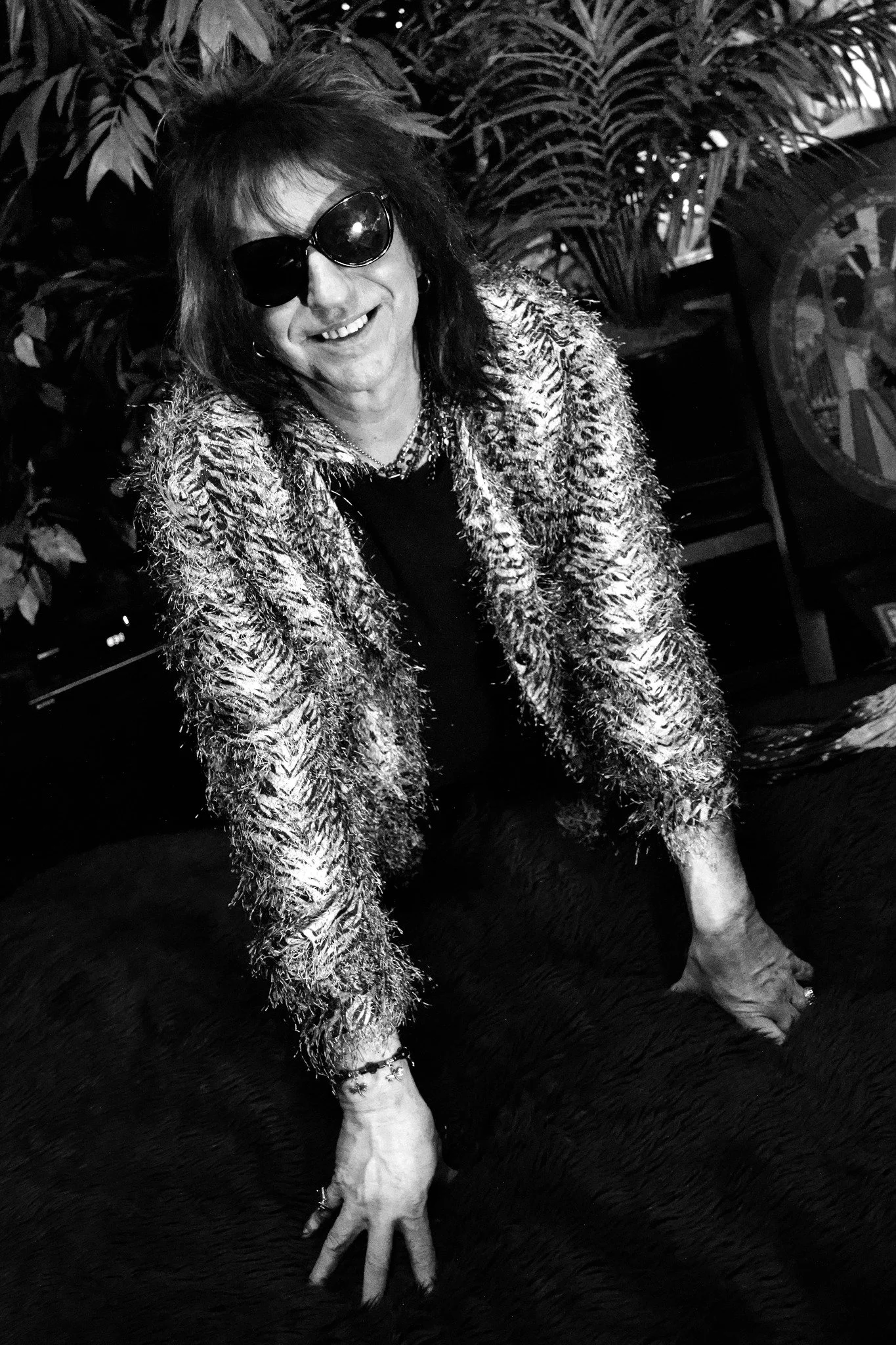 A person with dark hair, wearing sunglasses, a textured jacket, and jewelry, smiling and leaning on a large furry surface in an indoor setting with plants and a fan in the background.