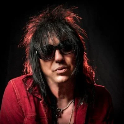 Person with spiky black hair wearing sunglasses and a red jacket, posing against a dark background.