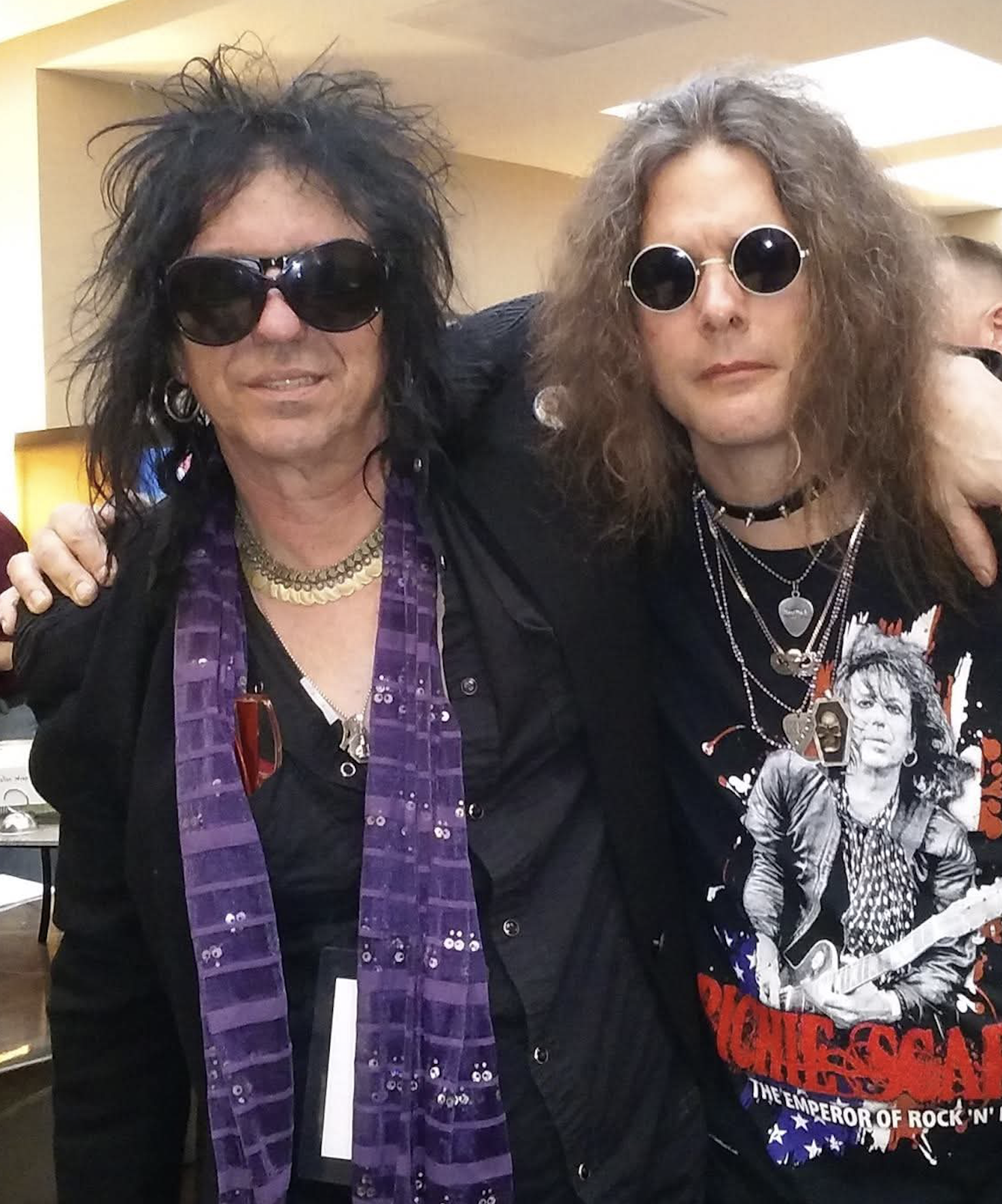 Two people with long, curly hair and sunglasses, dressed in rock-style clothing, posing together indoors.