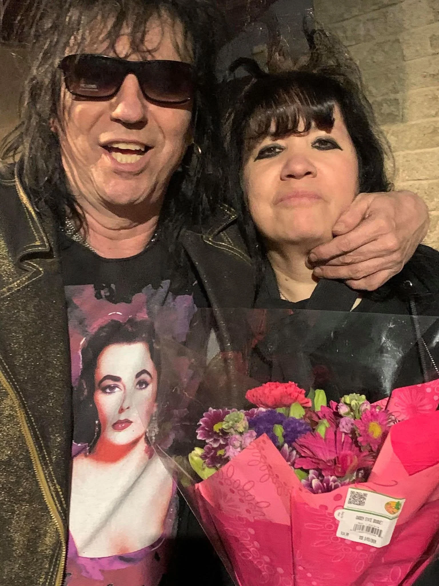 Two people, a man with sunglasses and long black hair, and a woman with short black hair and bangs, pose together. The woman is holding a bouquet of pink and purple flowers wrapped in pink paper, which features an image of a woman on it.