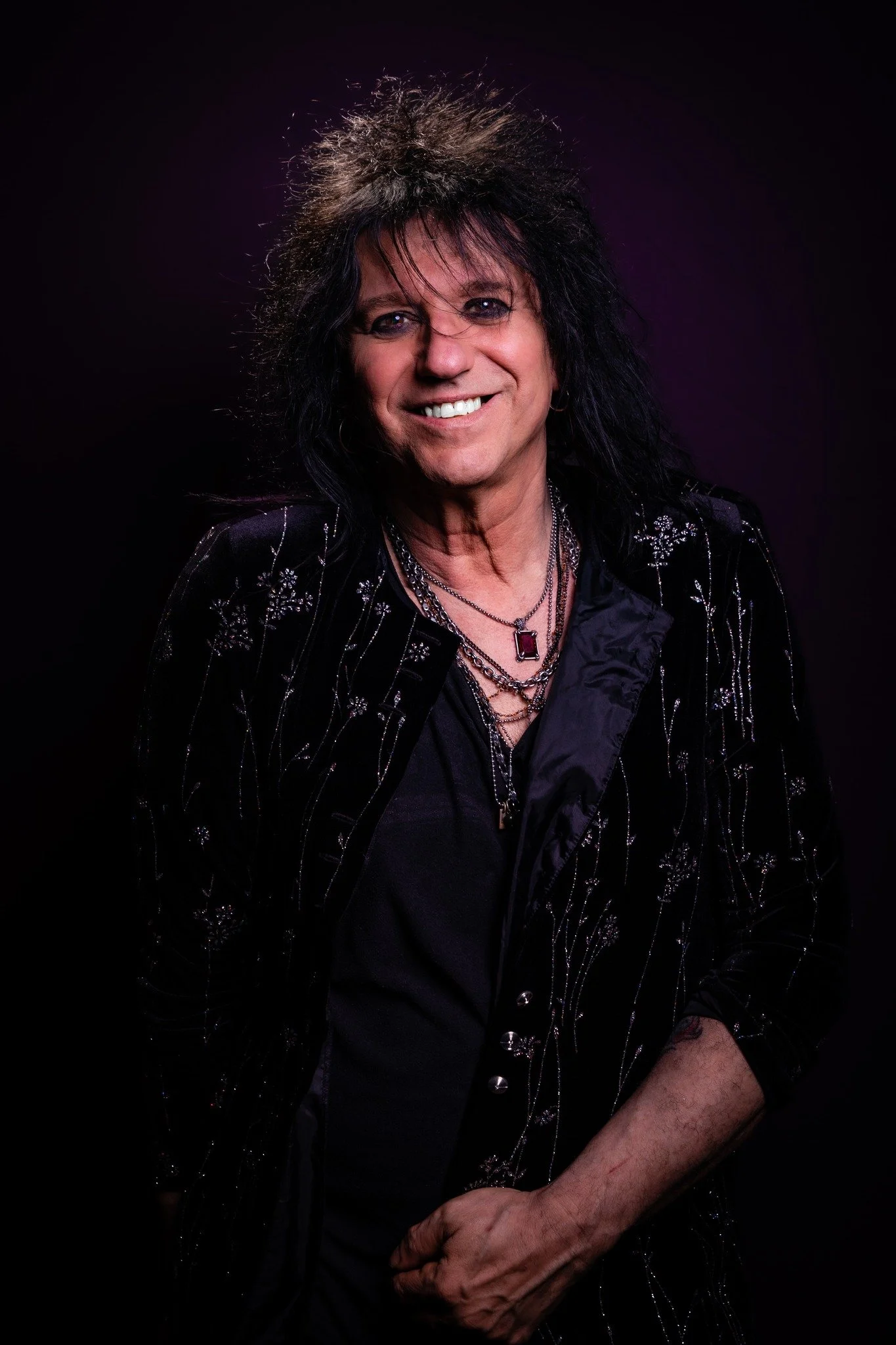 Portrait of a smiling man with long, dark, wavy hair, wearing a black jacket with silver embroidery and multiple layered necklaces, against a dark background.